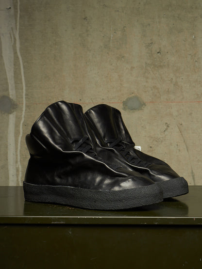 A pair of black leather high-top sneakers with sculpted, wrinkled textures and thick black soles, displayed on a dark green table against a textured concrete wall.