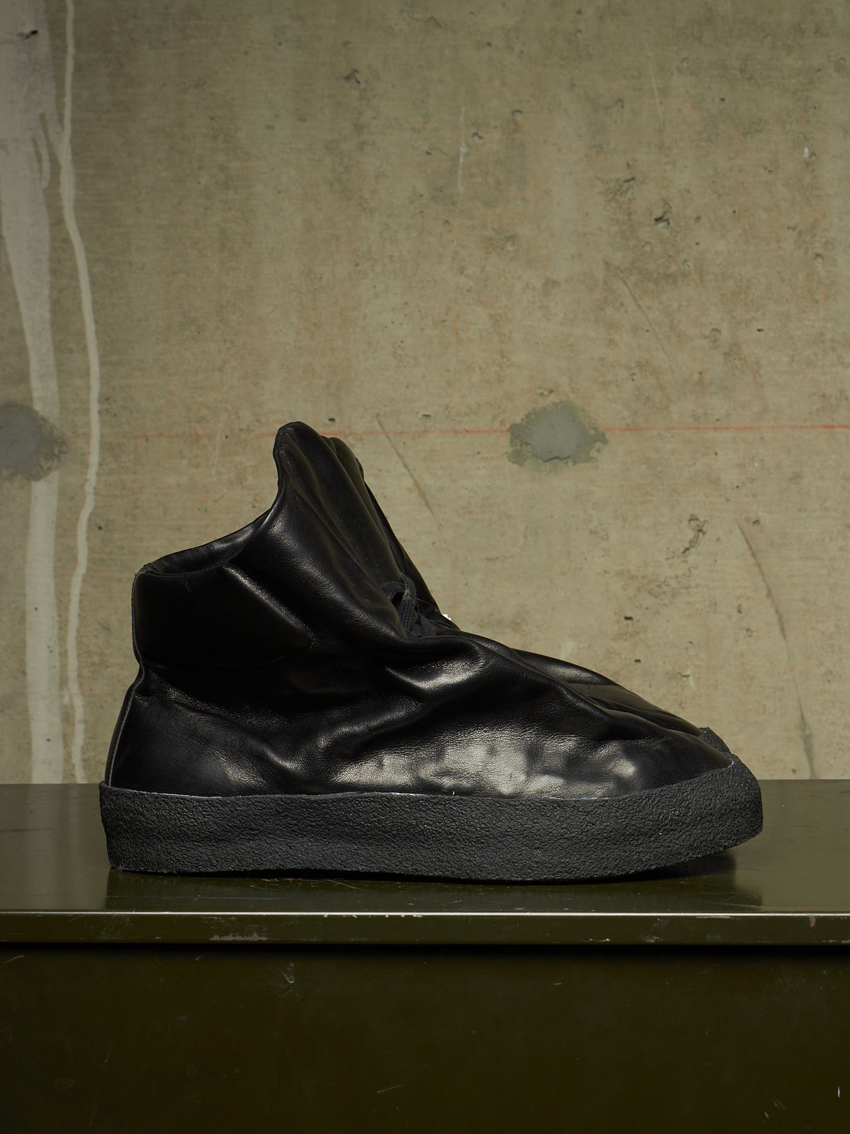 A black high-top sneaker with a wrinkled, slouchy design and a thick textured black sole, displayed on a dark surface against a textured concrete wall.