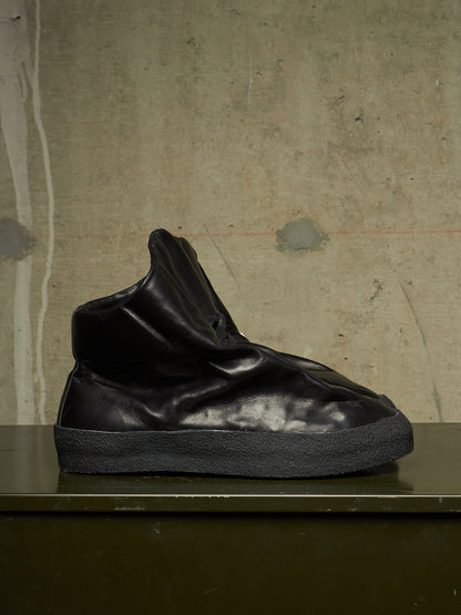 A black high-top sneaker with a wrinkled, slouchy design and a thick textured black sole, displayed on a dark surface against a textured concrete wall.
