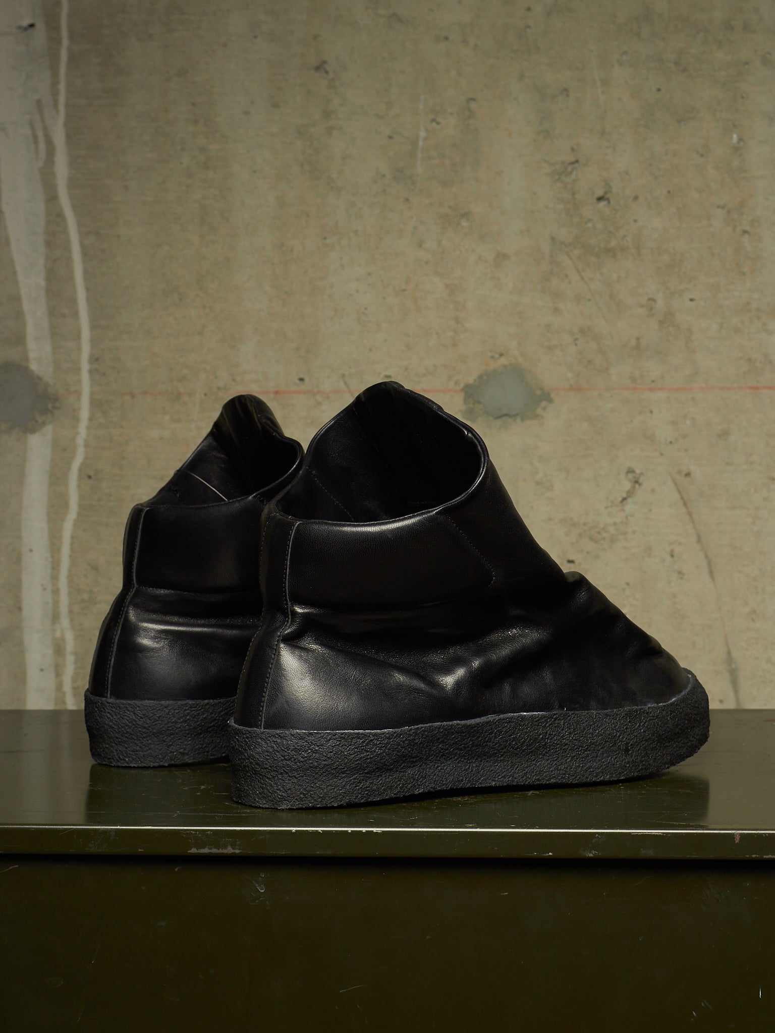 A pair of black leather high-top slip-on sneakers with thick black soles, displayed on a dark green shelf against a textured concrete wall.