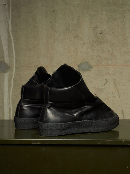 A pair of black leather high-top slip-on sneakers with thick black soles, displayed on a dark green shelf against a textured concrete wall.