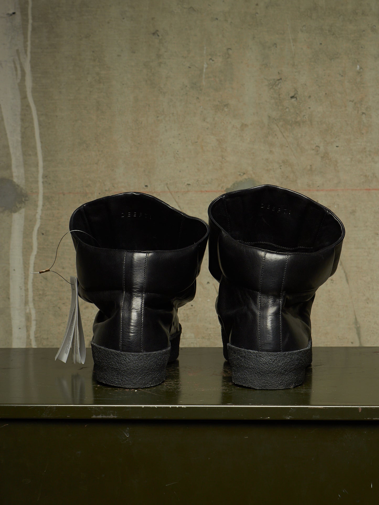 A pair of black leather ankle boots with chunky black soles, shown from the back on a dark green shelf against a textured concrete wall.