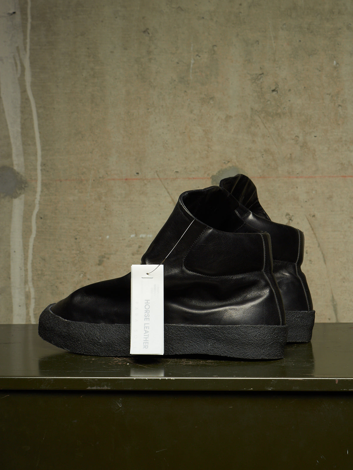 A pair of black leather Chelsea-style ankle boots with thick black soles, displayed on a dark green shelf against a textured concrete wall. A white hangtag is attached to the front boot.