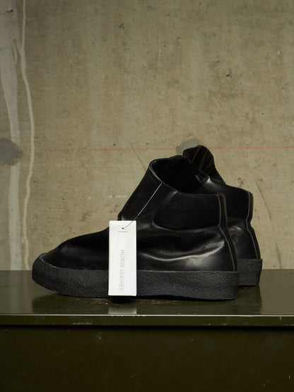 A pair of black leather Chelsea-style ankle boots with thick black soles, displayed on a dark green shelf against a textured concrete wall. A white hangtag is attached to the front boot.