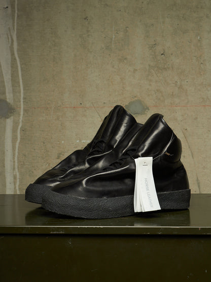 A pair of black leather high-top sneakers with thick black soles and white hang tags, displayed on a dark surface against a textured concrete wall.