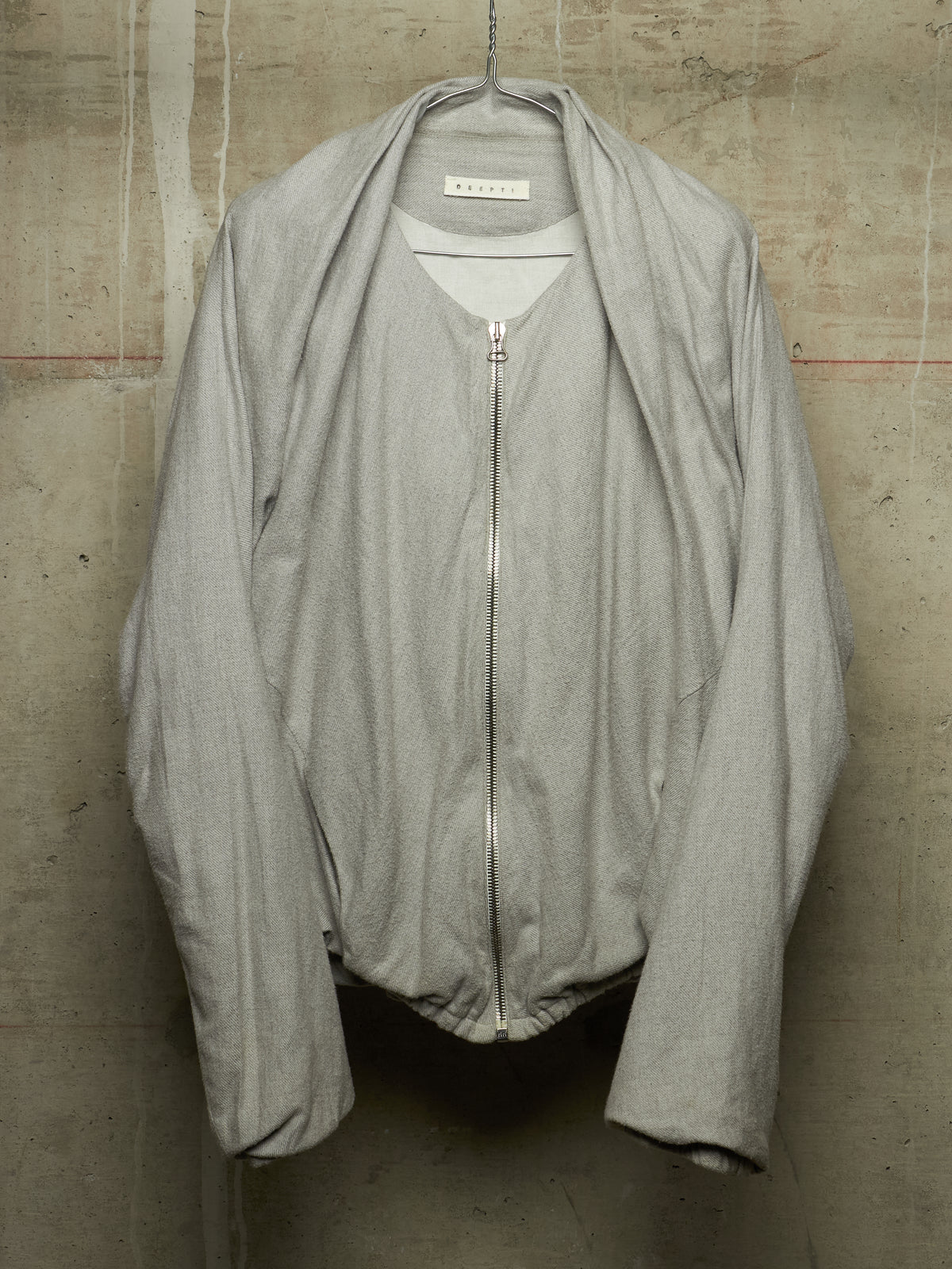 A light gray zip-up jacket with a wide collar and draped sleeves, hanging against a textured concrete wall. The brand label inside the collar reads 'M'ARMOUR'.