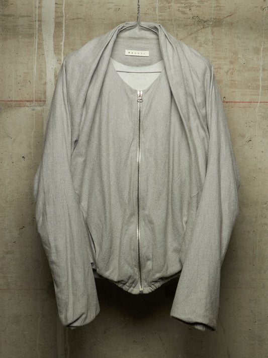 A light gray zip-up jacket with a wide collar and draped sleeves, hanging against a textured concrete wall. The brand label inside the collar reads 'M'ARMOUR'.