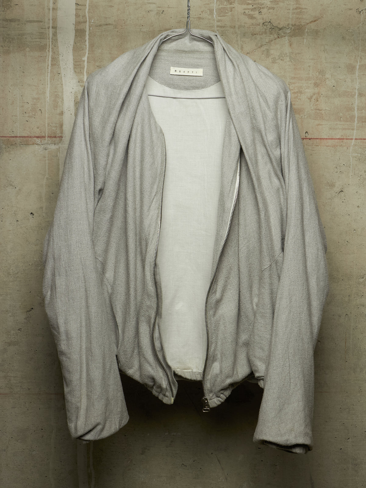 A light gray open-front cardigan or lightweight jacket draped over a white shirt, hanging against a textured concrete wall. The inner neck label shows the brand 'KUROO'.