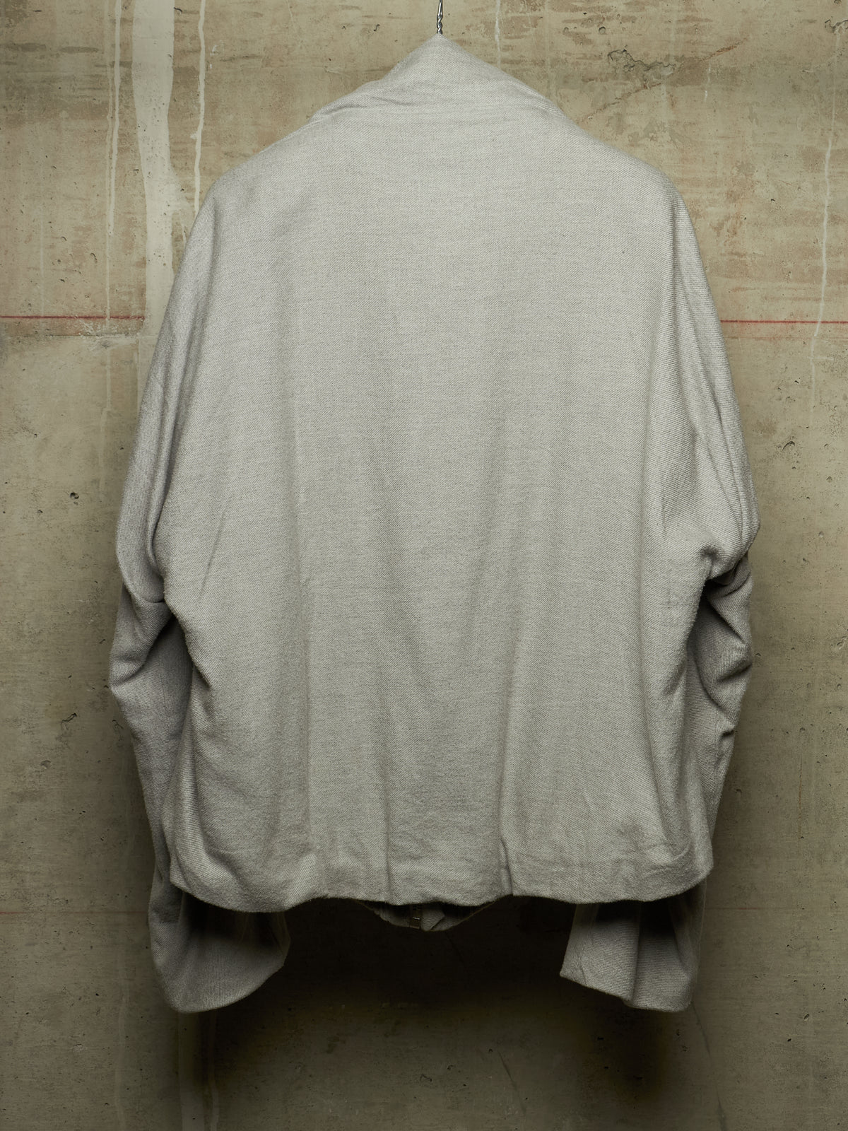 A light gray linen shirt jacket hanging against a textured concrete wall, showing the back view.