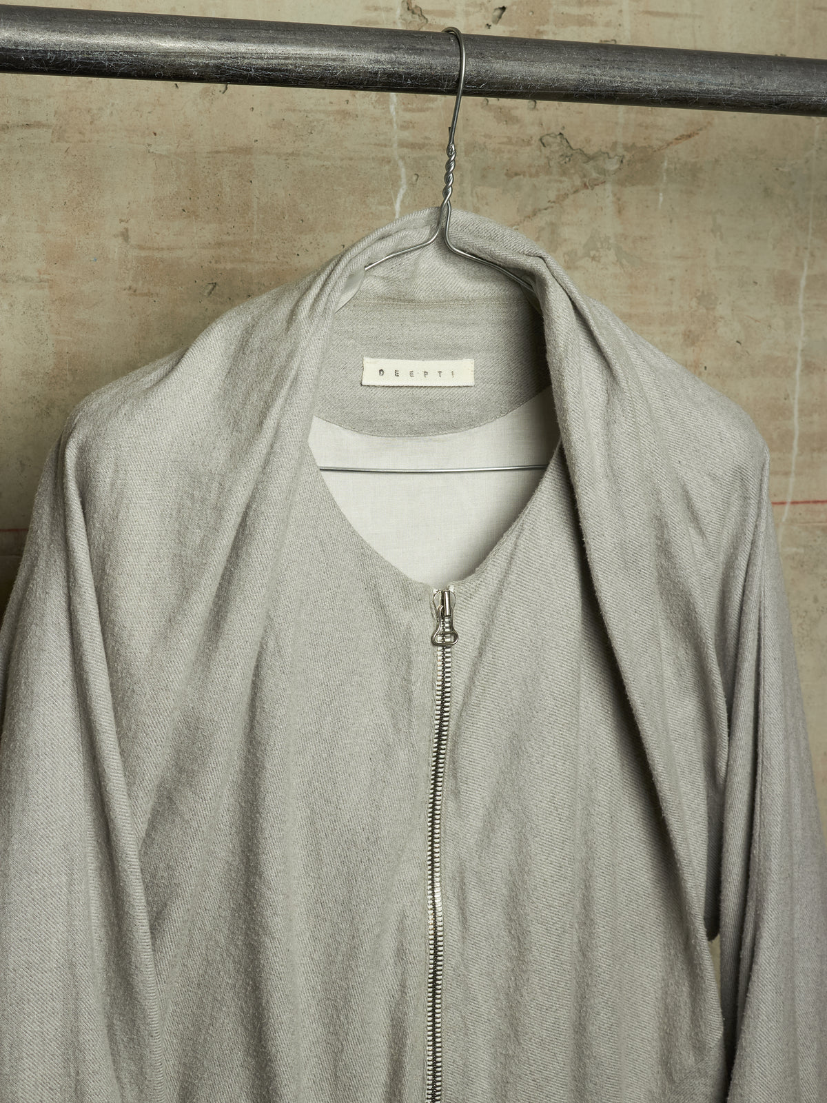 A light gray zip-up jacket with an oversized draped collar, hanging on a metal hanger from a dark metal rod against a textured beige wall. The brand name 'BERRY' is visible on the inner neck label.