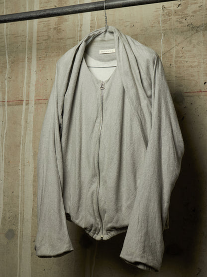 A light gray zip-up jacket with a draped collar, hanging on a metal hanger against a textured concrete wall.