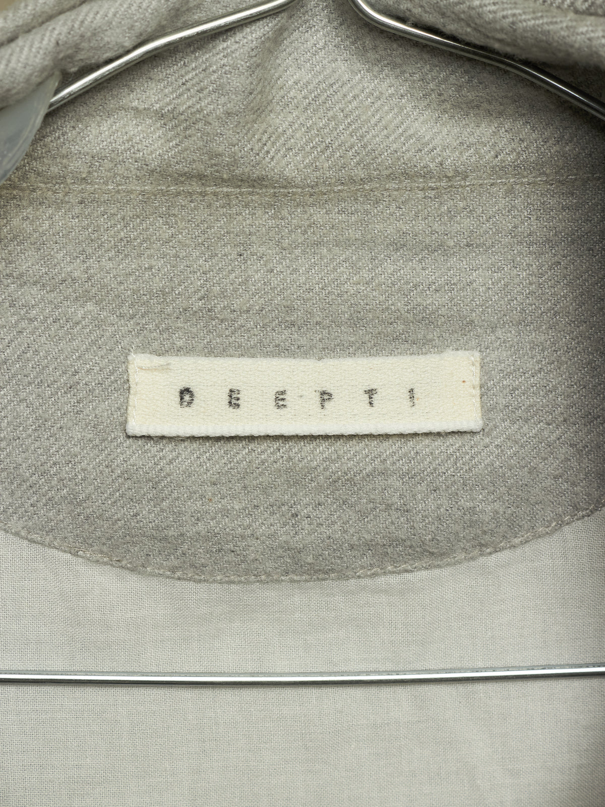 Close-up of the inside collar of a light gray woven garment on a metal hanger, showing a white fabric brand label that reads 'DEEPTI'.