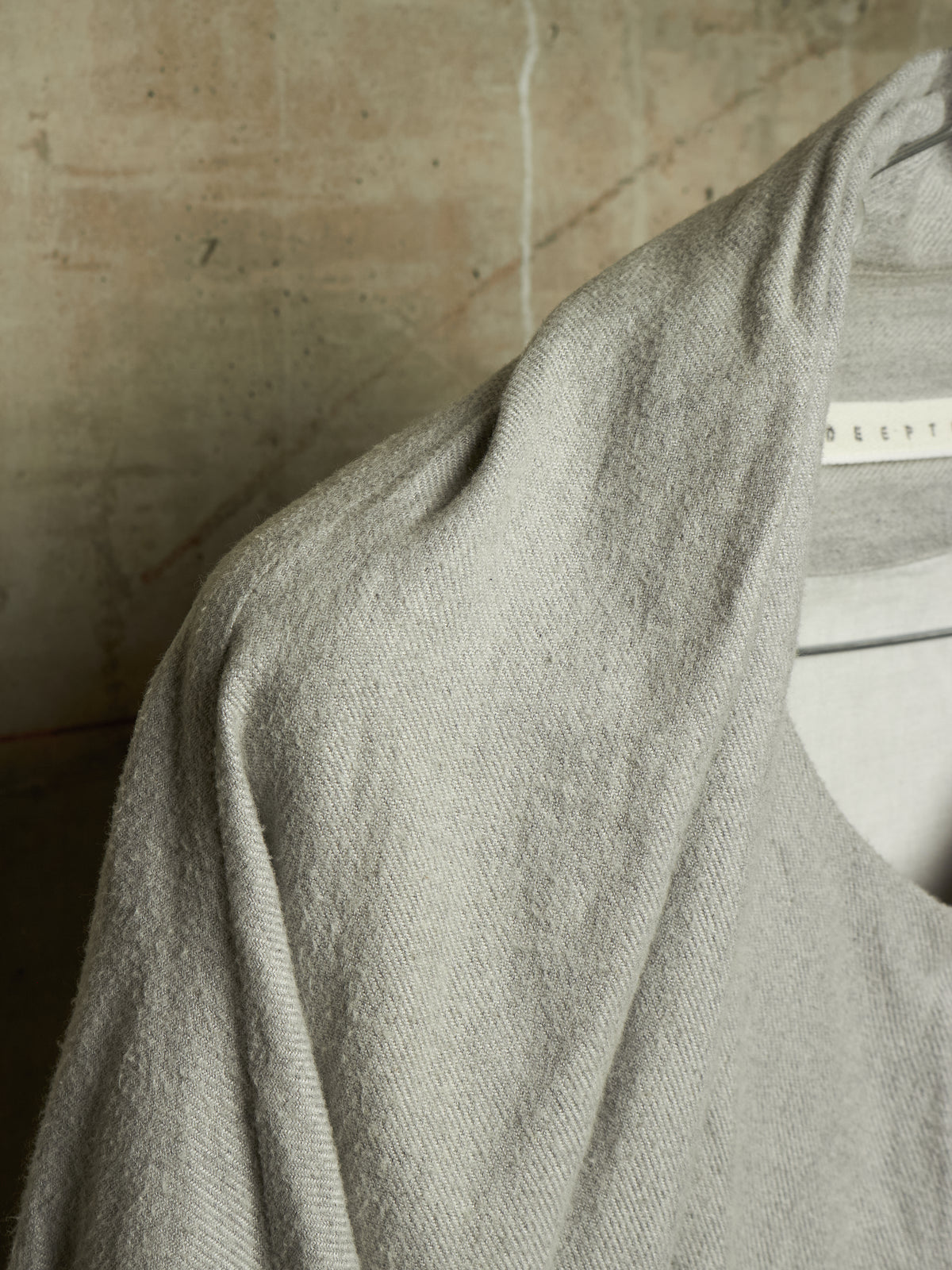 A close-up of a light gray knit garment draped over a white hanger, showing the soft texture and fine weave of the fabric against a neutral beige background.