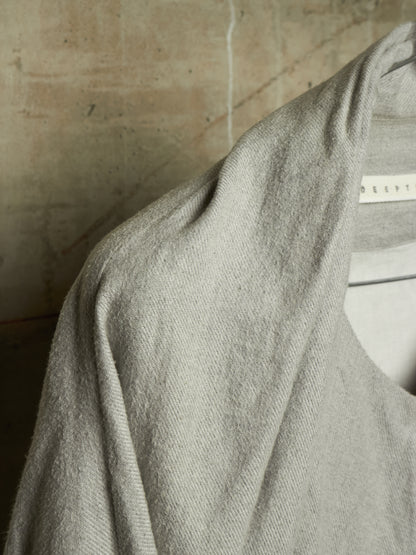 A close-up of a light gray knit garment draped over a white hanger, showing the soft texture and fine weave of the fabric against a neutral beige background.