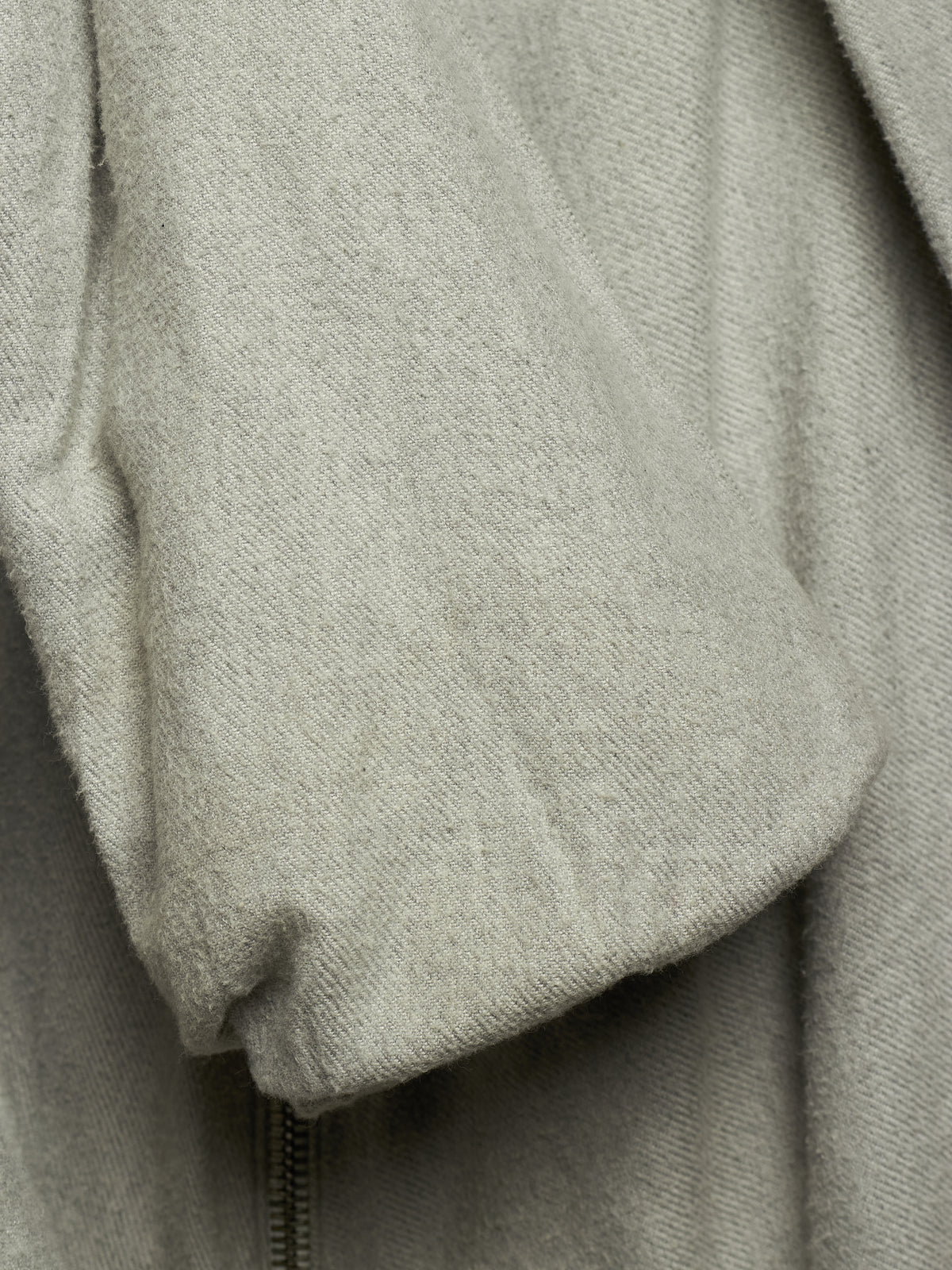 Close-up of a light gray textured fabric garment, showing a soft draped sleeve or cuff with a zipper detail at the bottom.