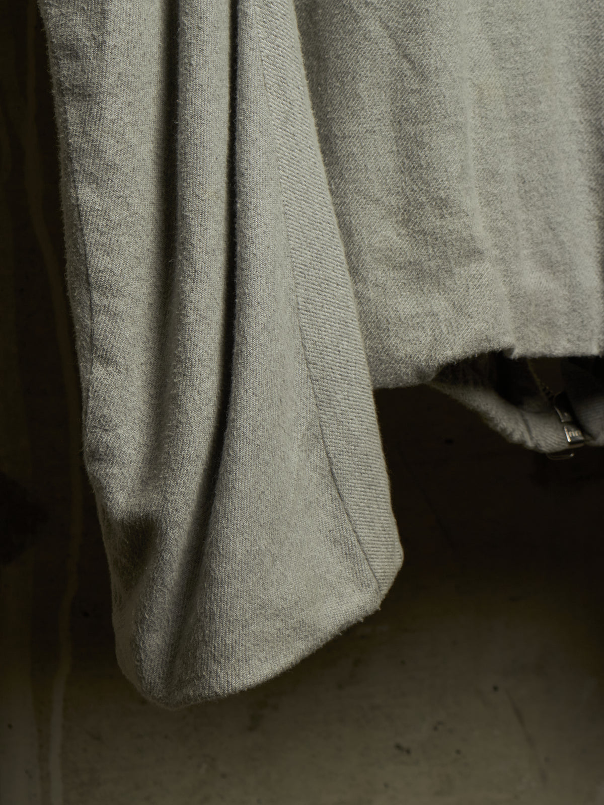 Close-up of a light gray textured fabric garment, showing a folded cuff or hem with a visible metal zipper pull against a dark background.