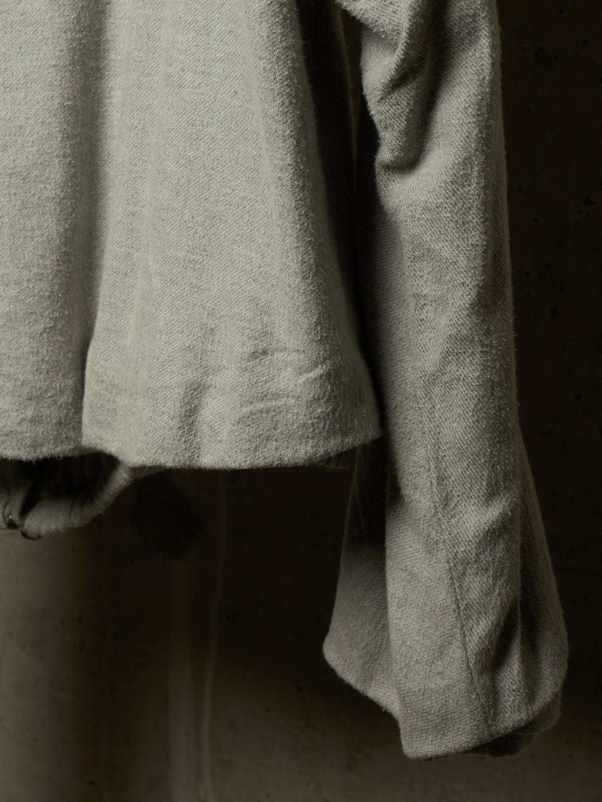 A close-up of a light gray textured fabric garment, showing the soft weave and draped folds against a dark, neutral background.