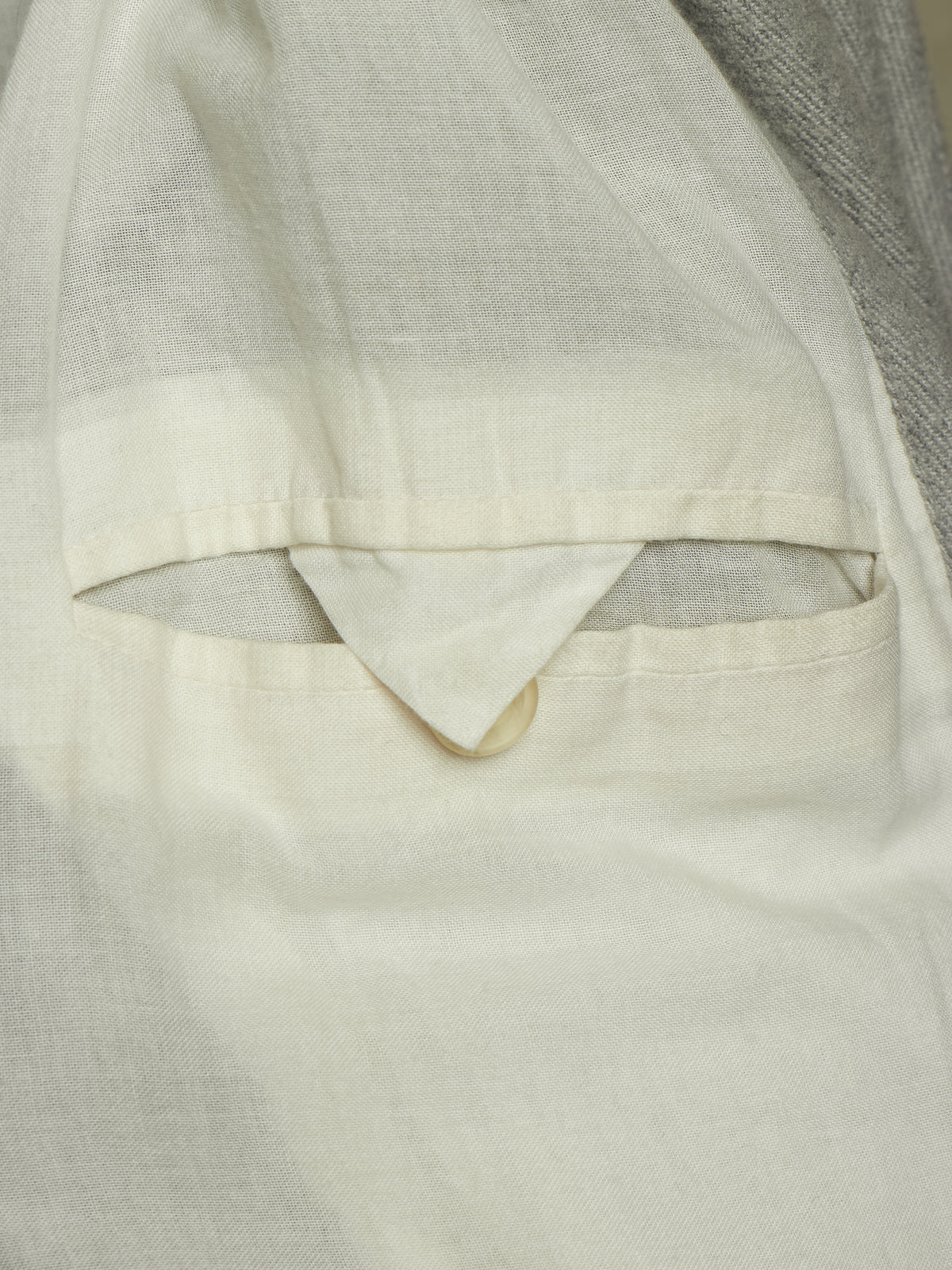 Close-up detail of a light cream or off-white linen garment, showing a pocket opening with a folded triangular flap and a small button, against a neutral gray fabric background.