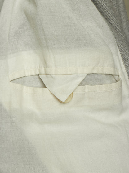 Close-up detail of a light cream or off-white linen garment, showing a pocket opening with a folded triangular flap and a small button, against a neutral gray fabric background.