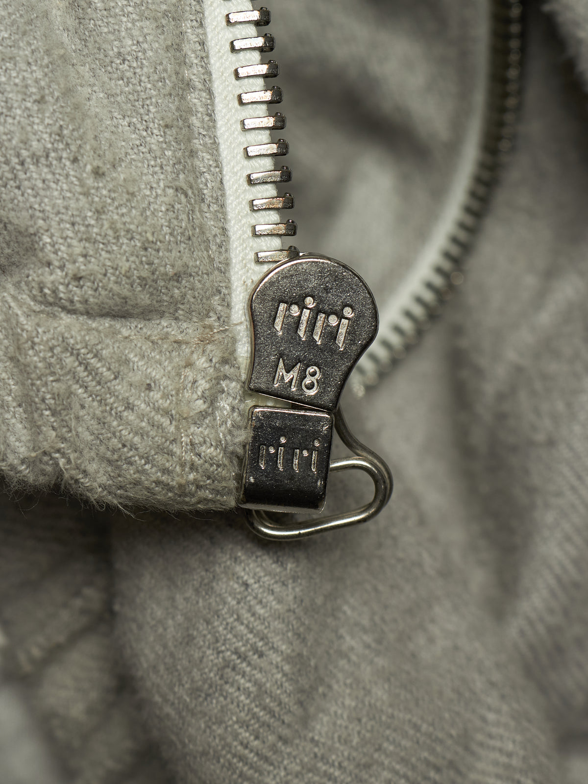 Close-up of a gray zippered garment with a silver metal zipper pull engraved with the brand name 'riri' and size 'M8'.