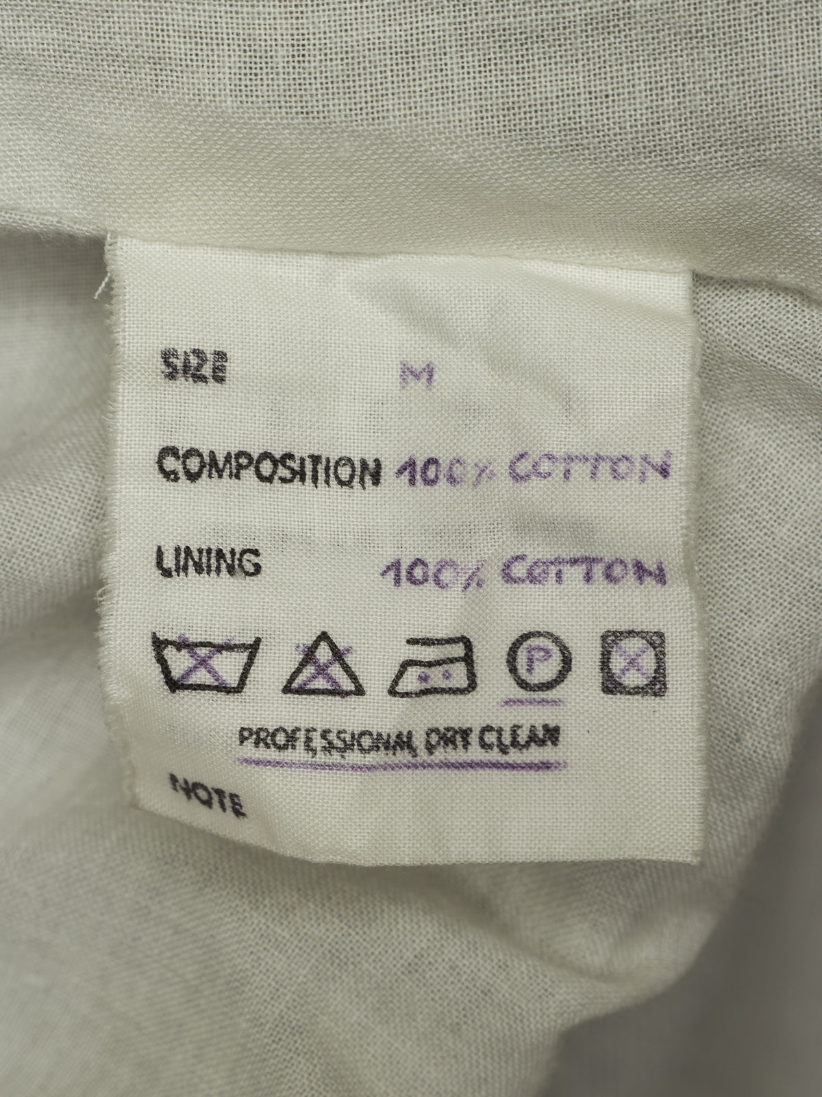 Close-up of a white fabric garment label showing size M, 100% cotton composition and lining, with care symbols and 'Professional Dry Clean' text.