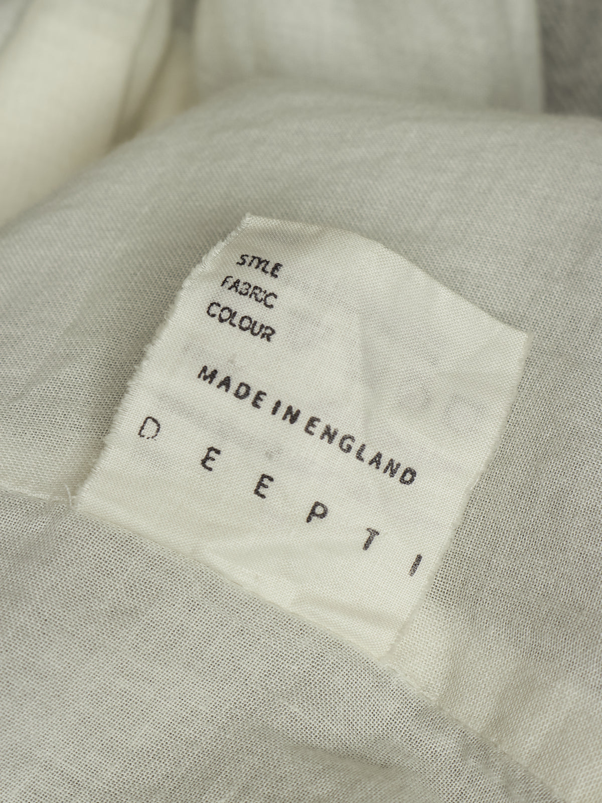 Close-up of a light beige fabric garment with a white sewn-in label reading 'DEEPTI MADE IN ENGLAND' and 'STYLE FABRIC COLOUR'.