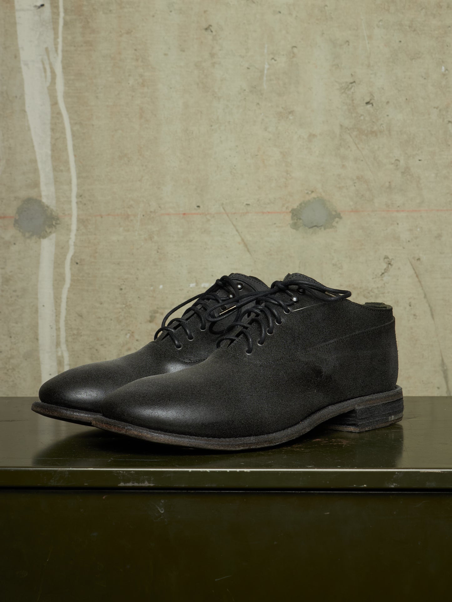A pair of black leather lace-up shoes with a chunky sole, displayed on a dark green surface against a textured beige concrete wall.