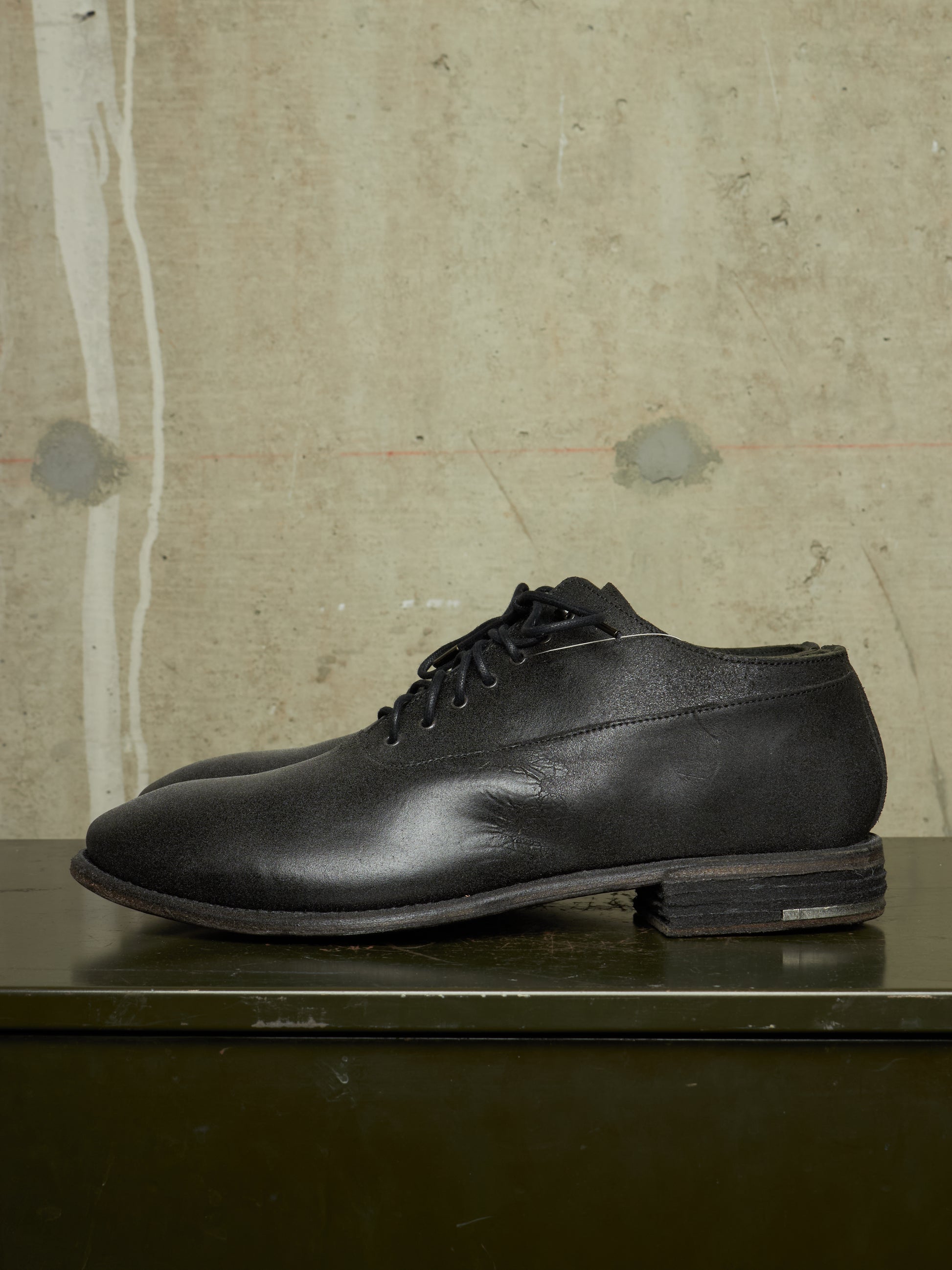 A single black leather lace-up dress shoe with a low stacked heel, resting on a dark green surface against a textured concrete wall background.