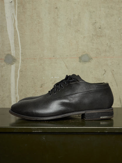 A single black leather lace-up dress shoe with a low stacked heel, resting on a dark green surface against a textured concrete wall background.