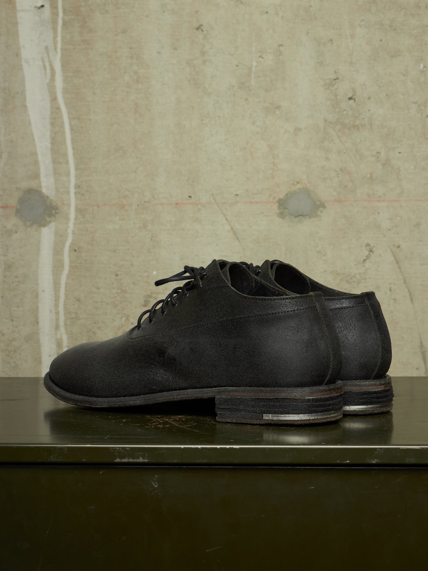 A pair of black leather lace-up shoes with a stacked lug sole, displayed on a dark green surface against a textured concrete wall.