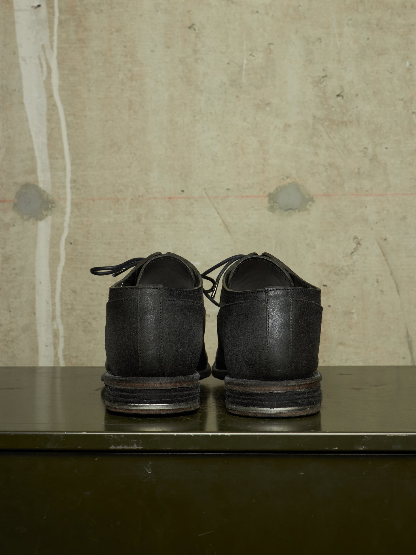 A pair of black leather lace-up shoes shown from the back, resting on a dark green metal surface against a textured beige concrete wall.