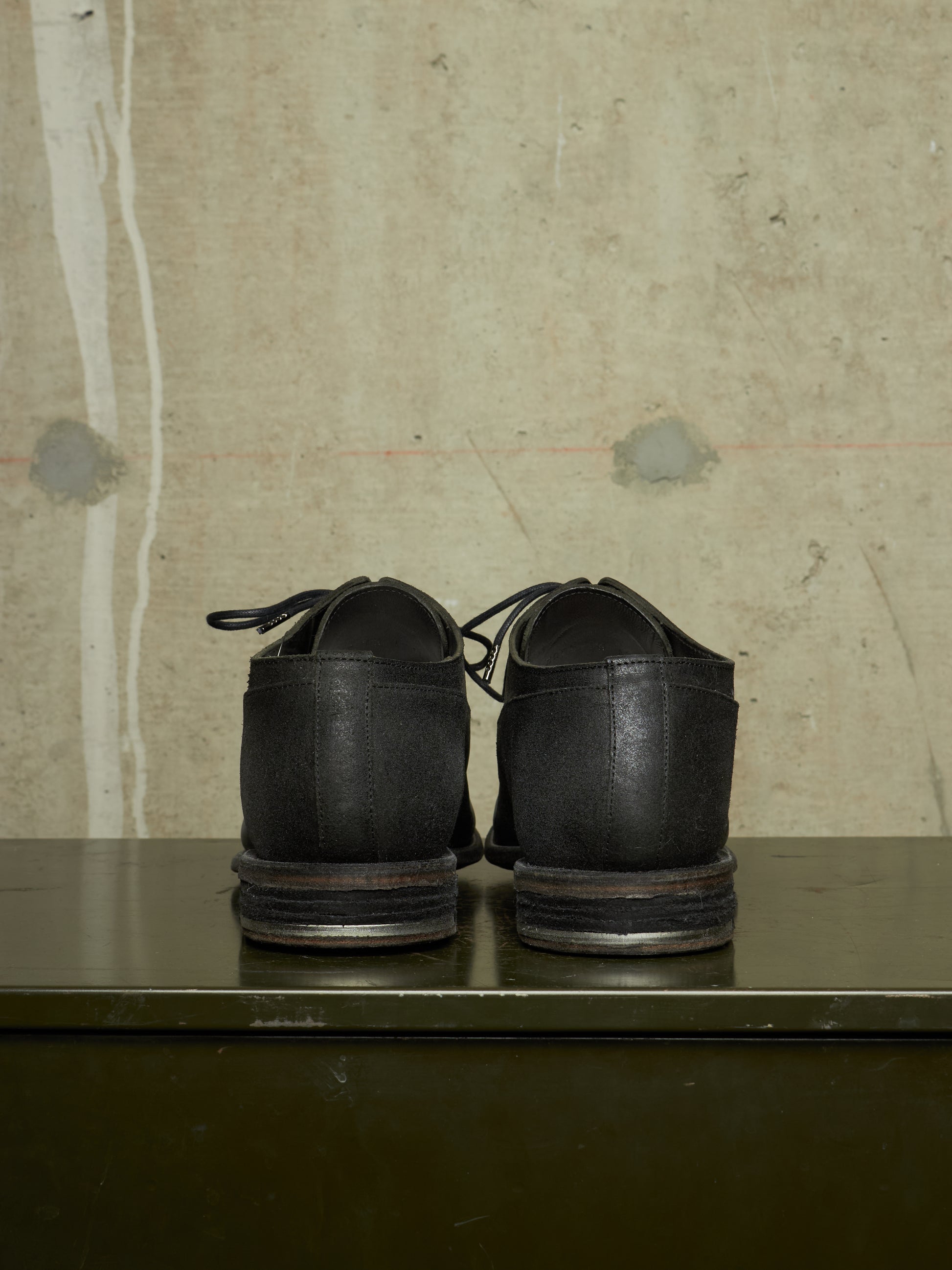 A pair of black leather lace-up shoes shown from the back, resting on a dark green metal surface against a textured beige concrete wall.
