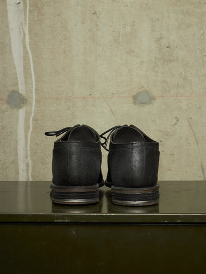 A pair of black leather lace-up shoes shown from the back, resting on a dark green metal surface against a textured beige concrete wall.