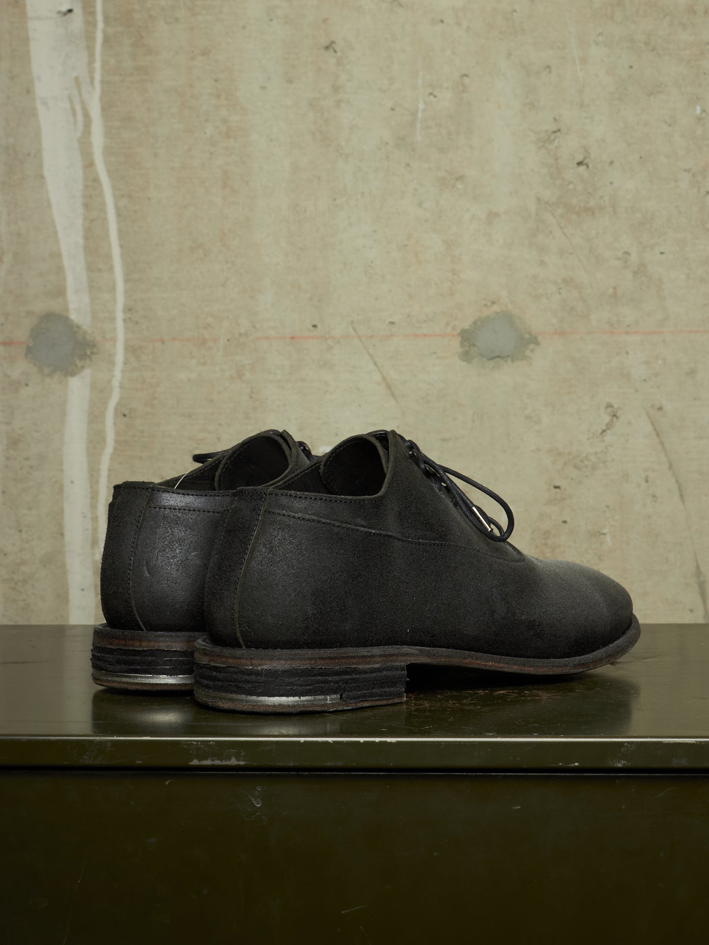 A pair of black leather lace-up dress shoes with stacked lug soles, displayed on a dark green metal surface against a textured beige concrete wall.