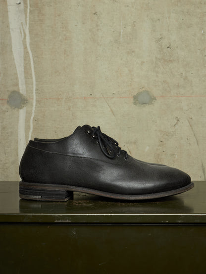 A black leather lace-up dress shoe with a low stacked heel, displayed on a dark green surface against a textured beige concrete wall.