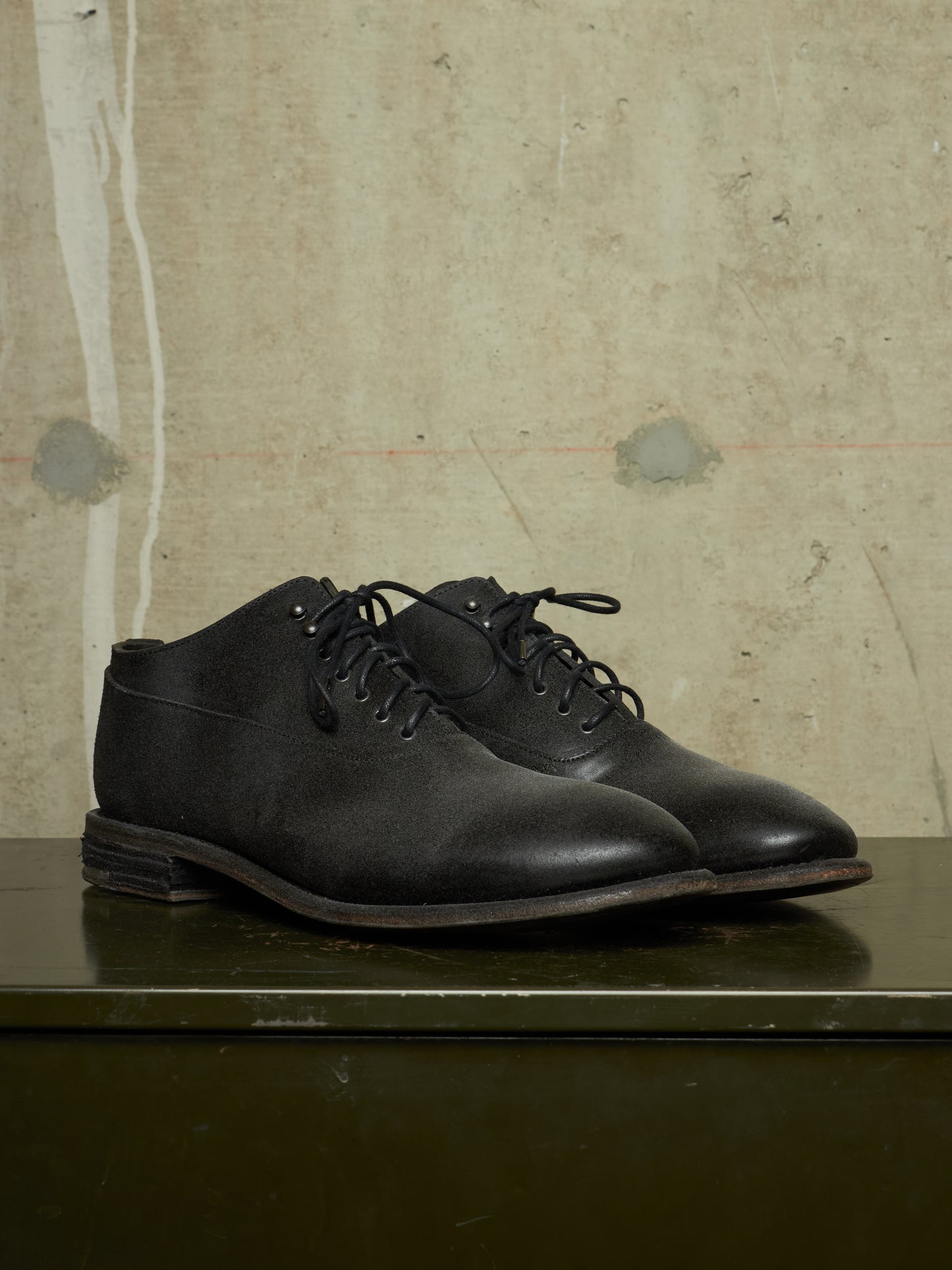 A pair of black leather lace-up ankle boots with a chunky sole, displayed on a dark green metal surface against a textured beige concrete wall.