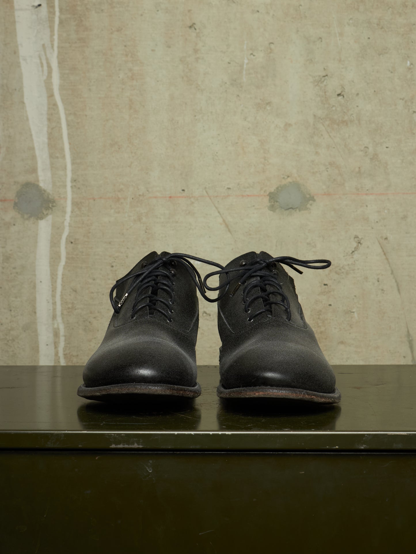 A pair of black leather lace-up dress shoes with a rounded toe, displayed on a dark green surface against a textured beige concrete wall.