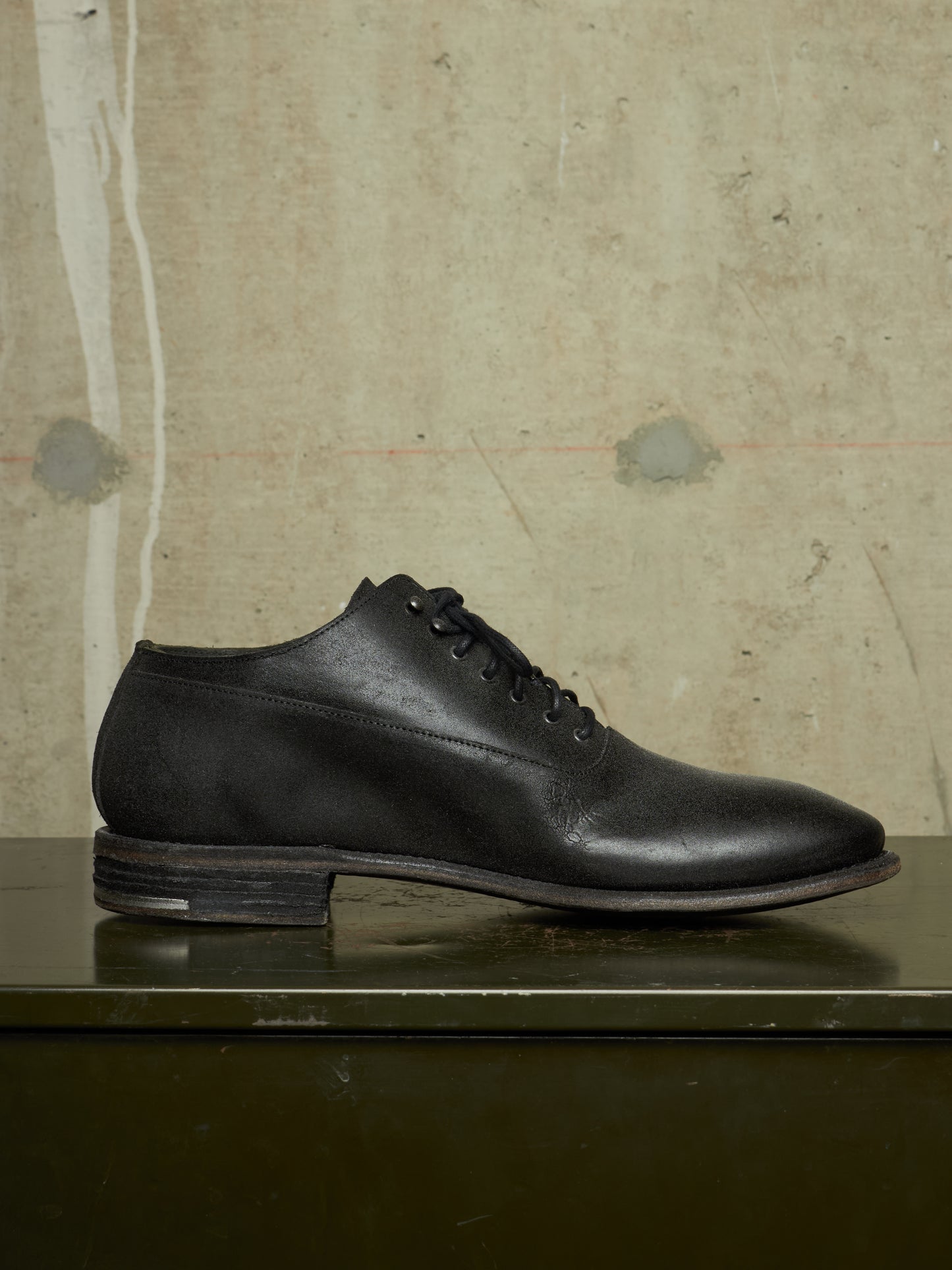 A single black leather lace-up dress shoe with a low stacked heel, displayed in profile on a dark reflective surface against a textured beige wall.
