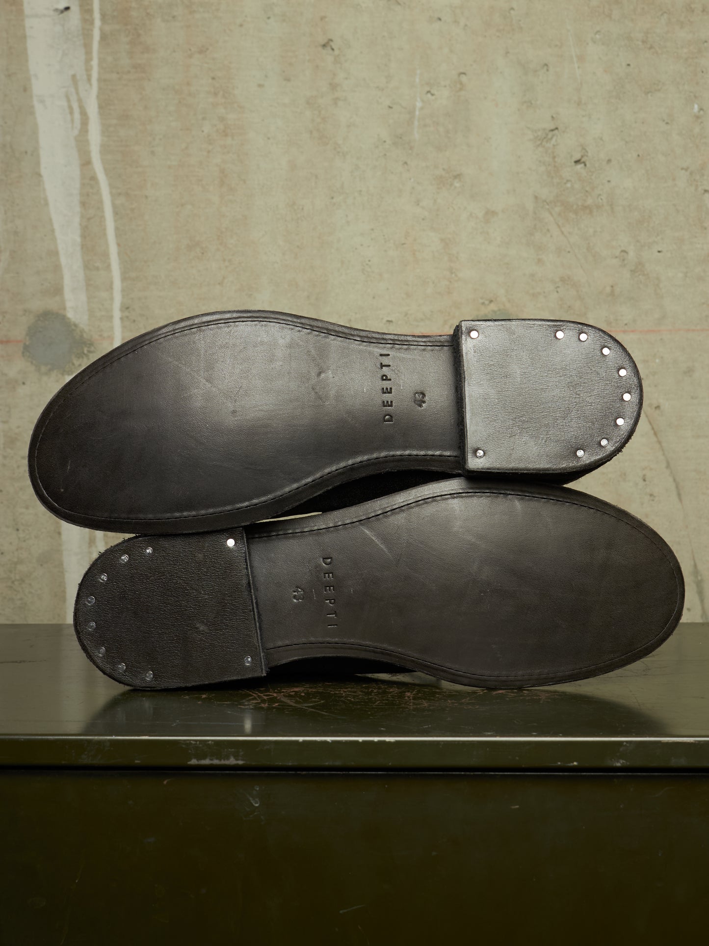 The soles of a pair of dark brown leather shoes, showing the 'GREAT STU' branding and decorative rivet details, resting on a dark surface against a textured beige background.