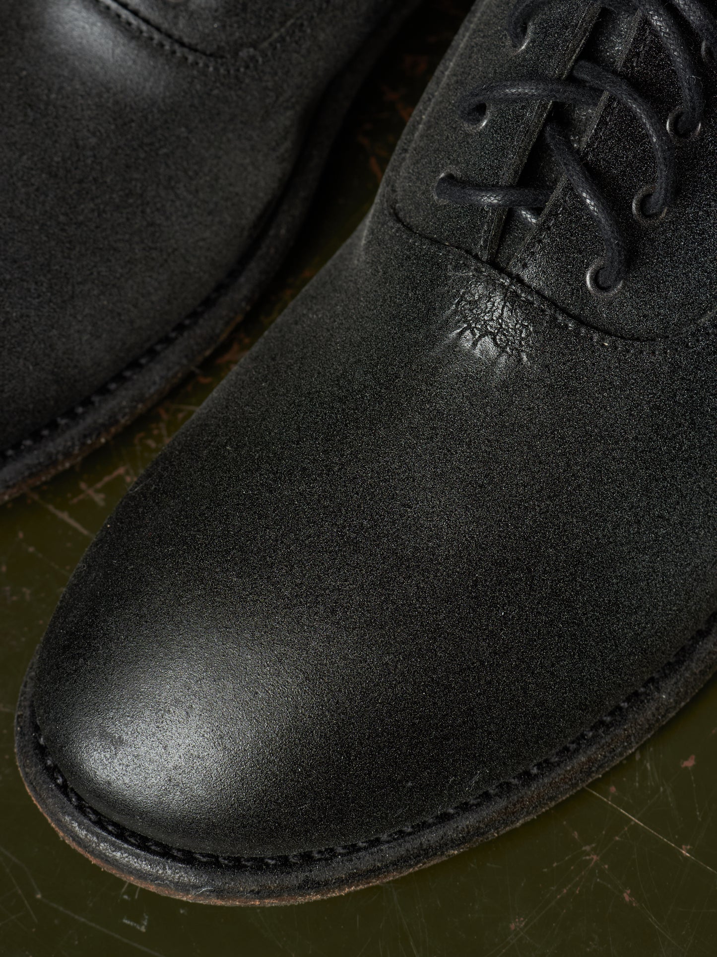 Close-up of a black leather lace-up boot with a rounded toe and visible stitching, resting on a dark olive-green surface.