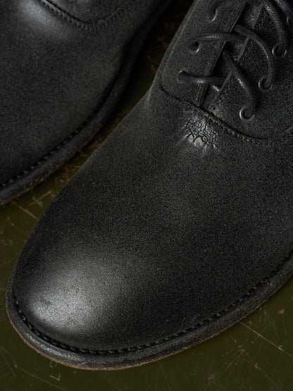Close-up of a black leather lace-up boot with a rounded toe and visible stitching, resting on a dark olive-green surface.