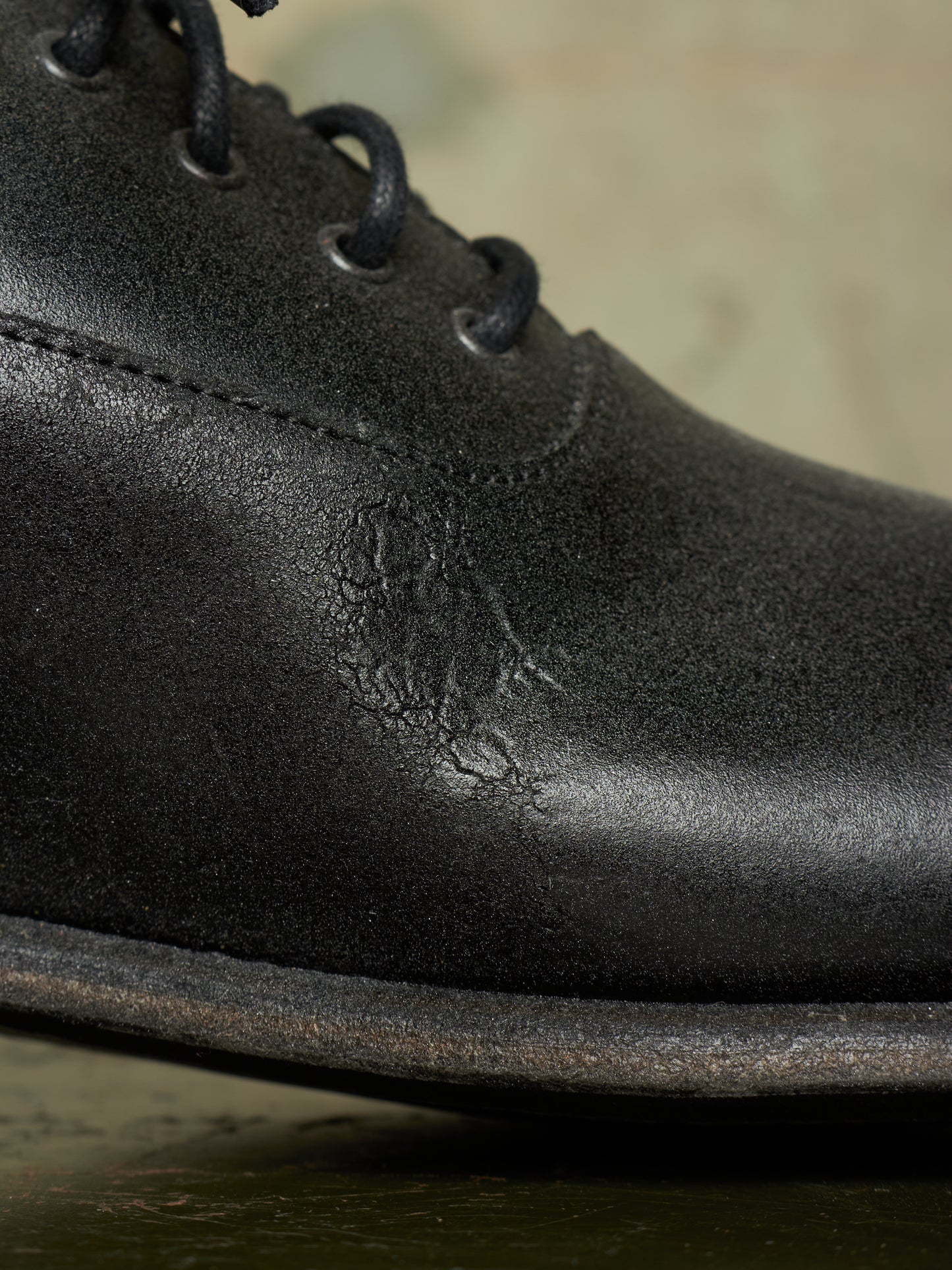 Close-up of a black leather lace-up boot showing the side panel, laces, and a subtle embossed logo on a textured background.