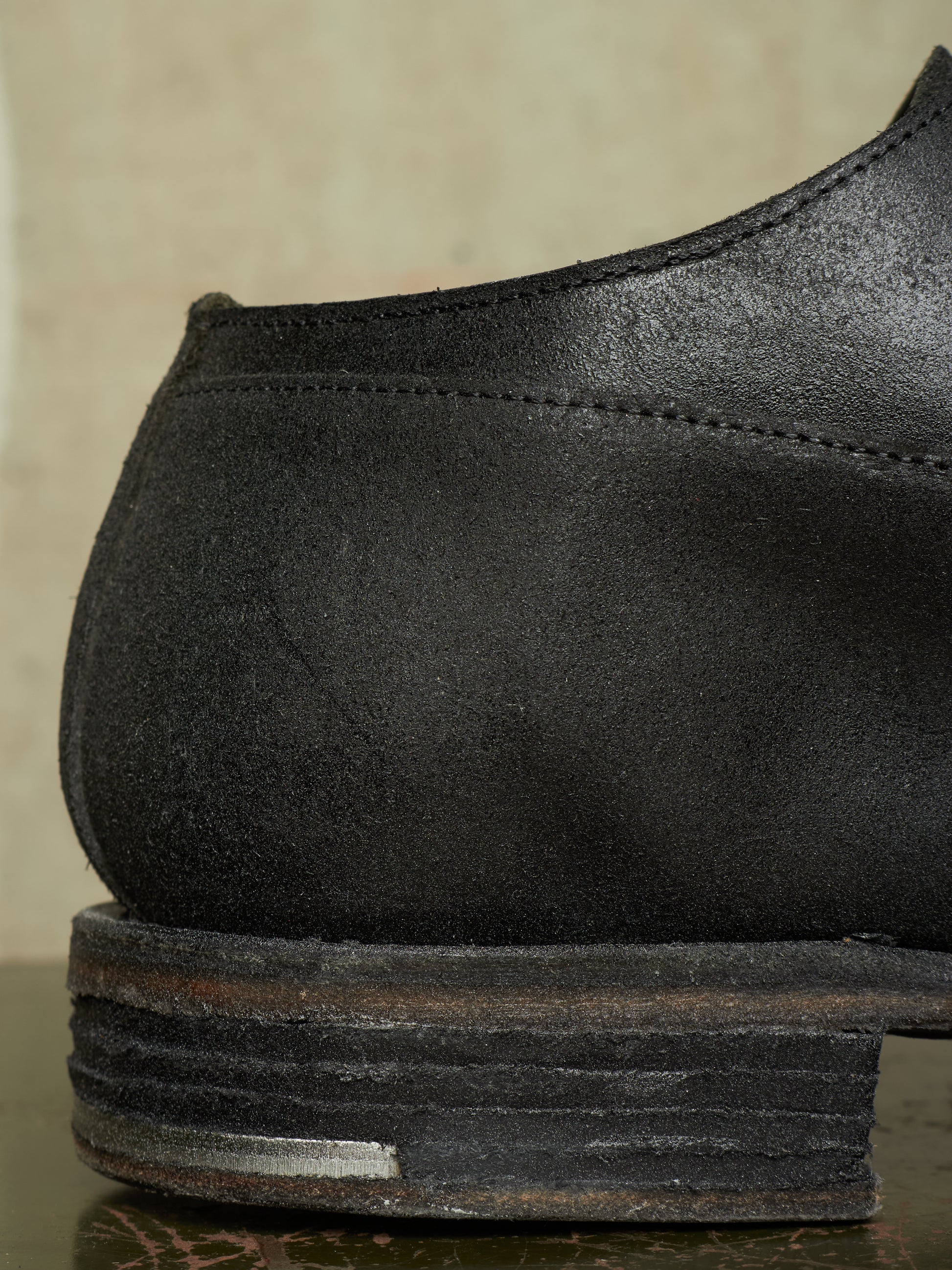 Close-up of the heel and stacked sole of a black leather shoe, showing the textured leather upper and weathered layered sole against a neutral beige background.