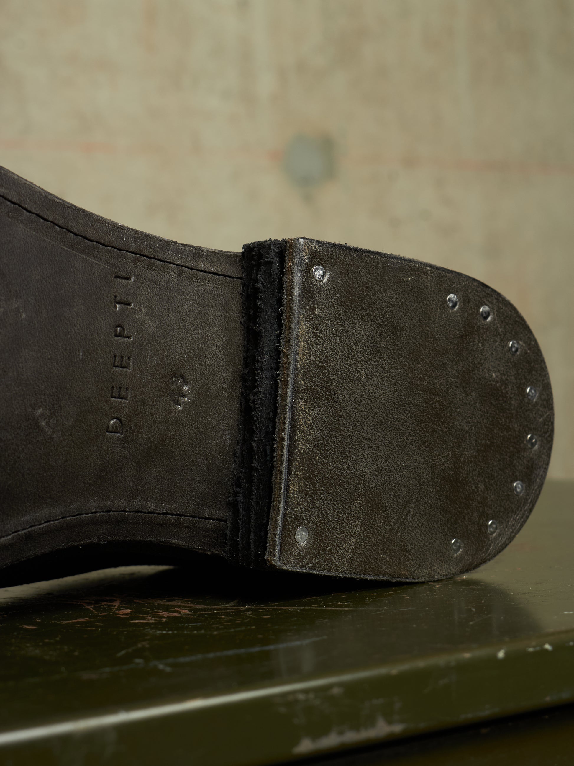 Close-up of the sole of a dark brown leather boot, showing the brand name 'DEEPTI' embossed on the heel area and metal rivets along the toe cap, resting on a dark green surface.