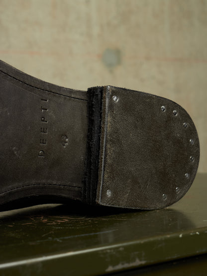 Close-up of the sole of a dark brown leather boot, showing the brand name 'DEEPTI' embossed on the heel area and metal rivets along the toe cap, resting on a dark green surface.