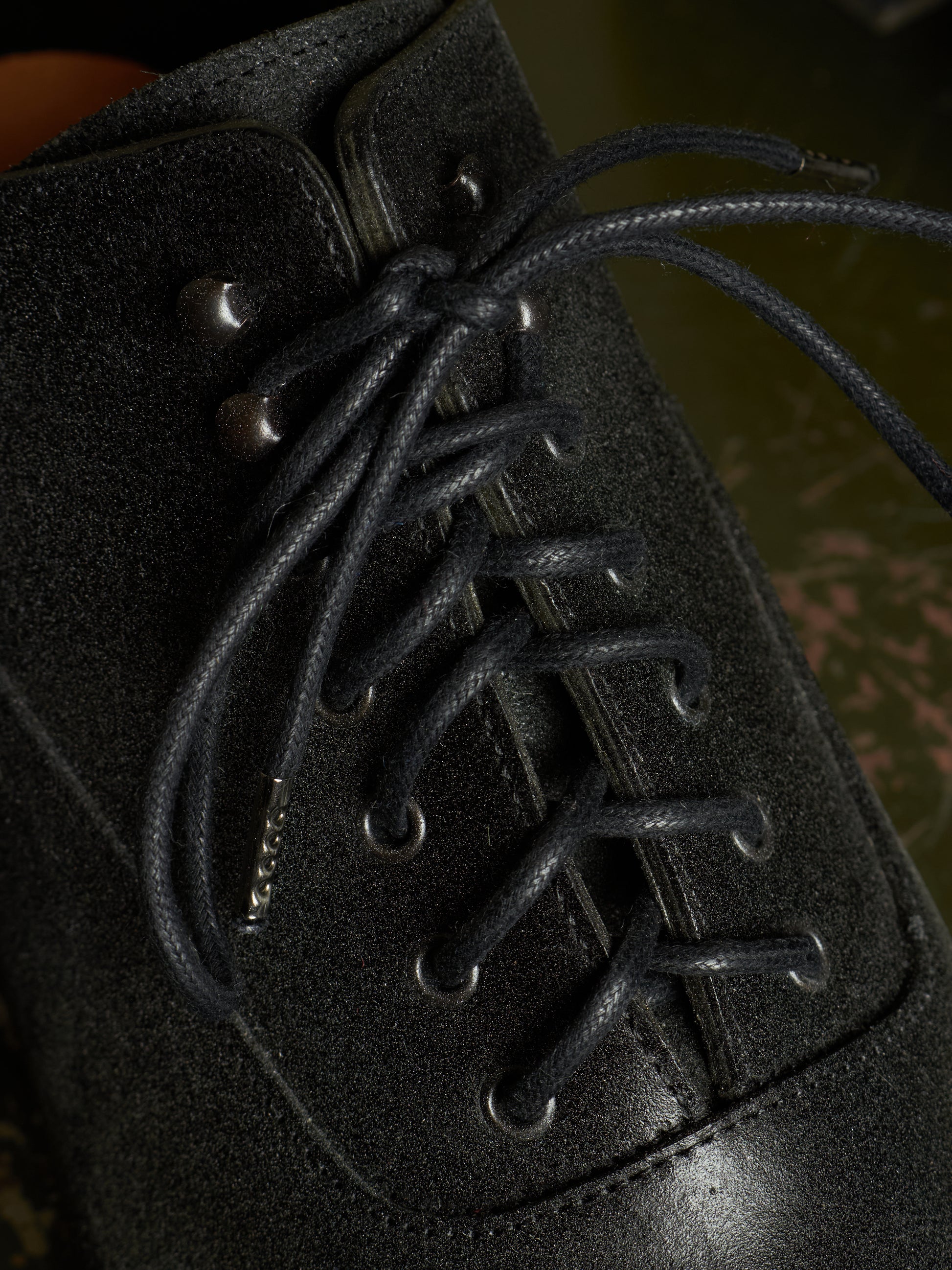 Close-up of a black suede lace-up boot with dark laces threaded through metal eyelets, set against a dark, blurred background.
