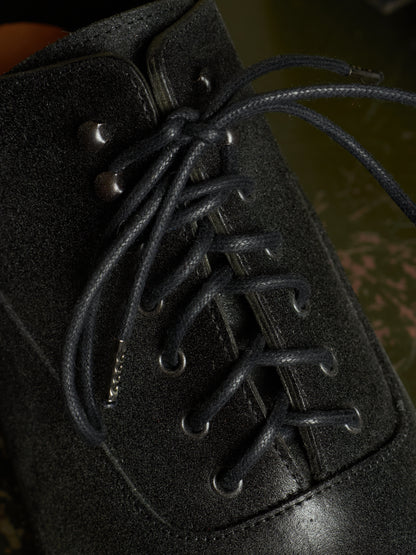 Close-up of a black suede lace-up boot with dark laces threaded through metal eyelets, set against a dark, blurred background.