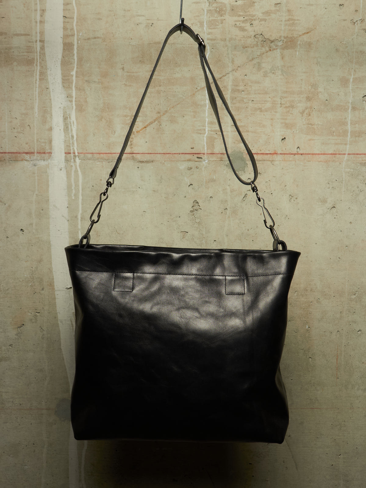 A black leather shoulder bag with a wide top and a long adjustable strap, hanging against a textured concrete wall.