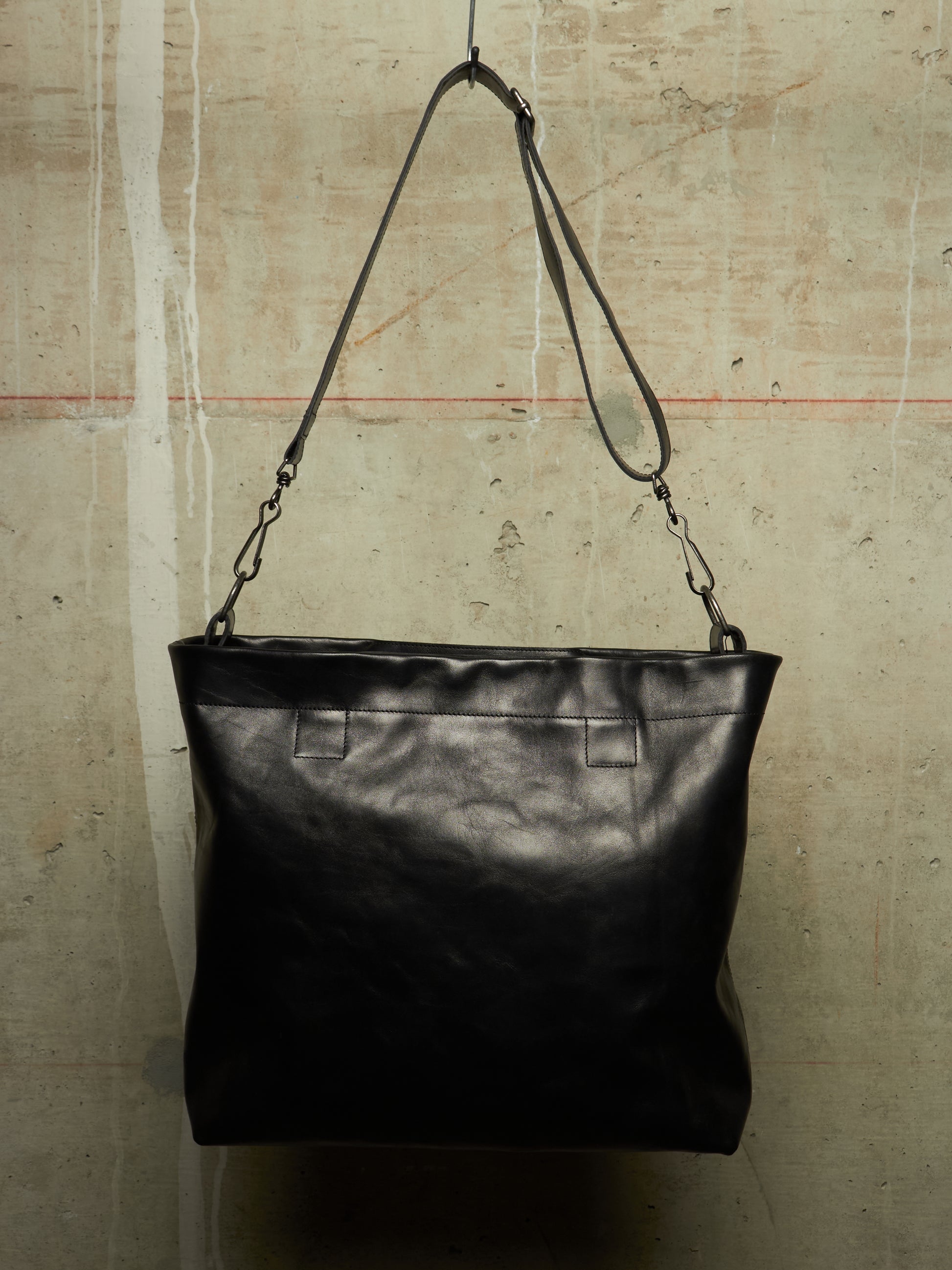 A black leather shoulder bag with a wide top and a long adjustable strap, hanging against a textured concrete wall.