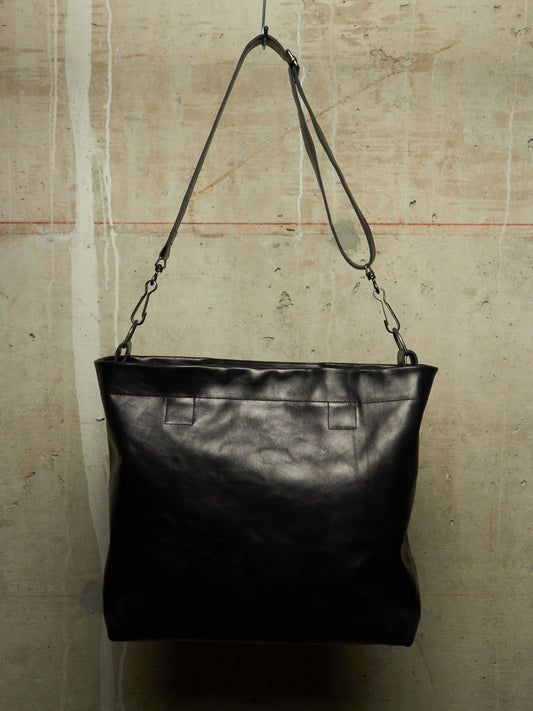 A black leather shoulder bag with a wide top and a long adjustable strap, hanging against a textured concrete wall.